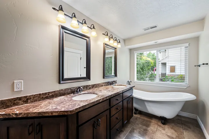 Spa-inspired master bathroom featuring a double vanity with rich wood cabinetry and engineered stone countertops, framed mirror with decorative sconces, freestanding soaking tub, and tile flooring throughout
