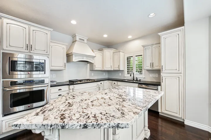 Kitchen featuring a large island with a stone countertop, built-in stainless steel appliances, custom cabinetry, a range hood, and dark wood-finish flooring