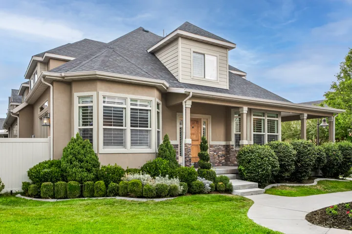 Stucco exterior with stone accents, covered porch with columns, dormer window, bay window with plantation shutters, and mature landscaping