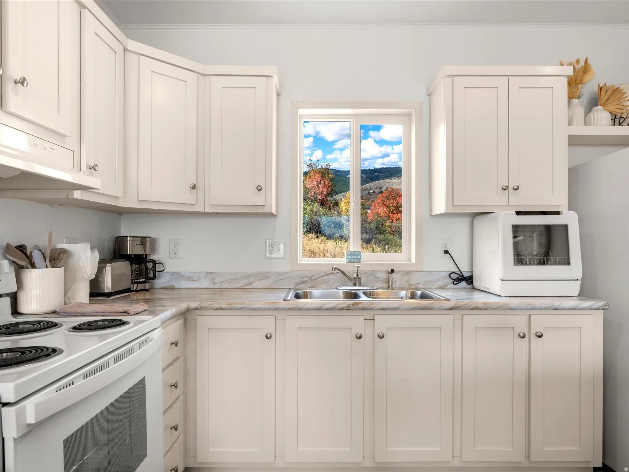 White cabinetry, neutral countertops, stainless steel double basin sink, electric range, and a window with a natural backdrop