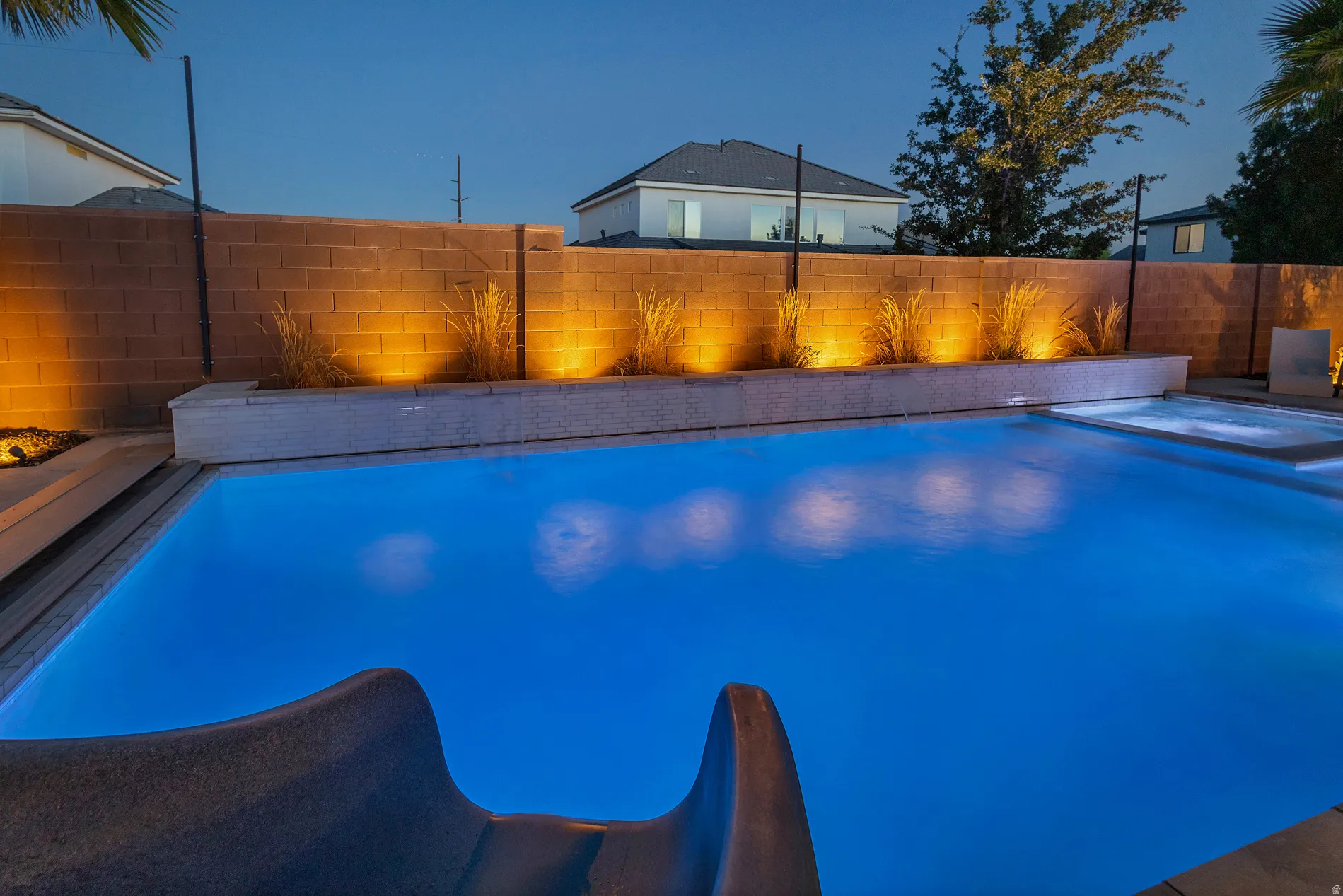Swimming pool with an integrated spa, a built-in water slide, perimeter accent lighting, and a textured privacy wall