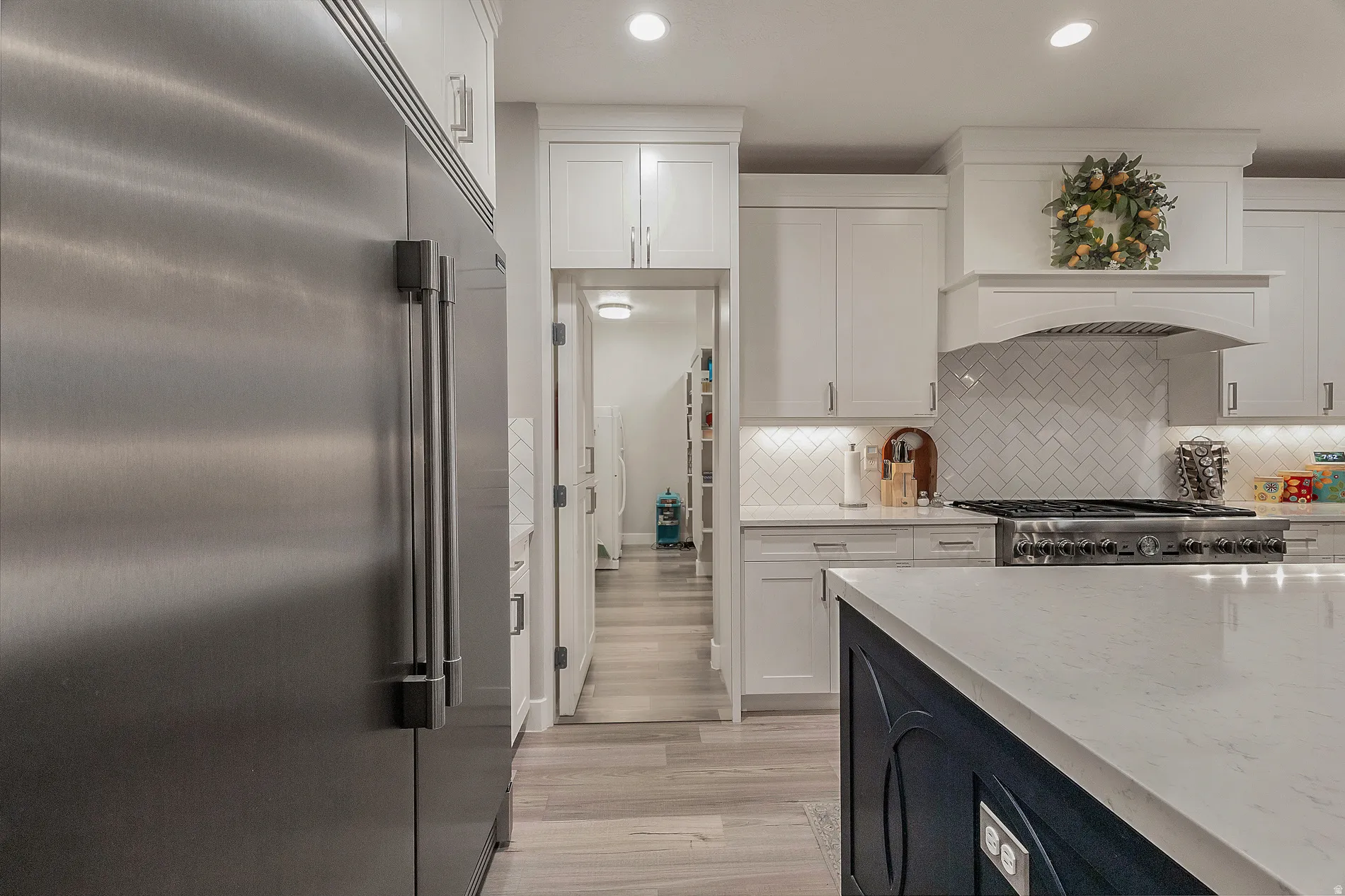 Stainless steel refrigerator, white shaker-style cabinetry, herringbone tile backsplash, range hood, and light-toned wood-finish flooring