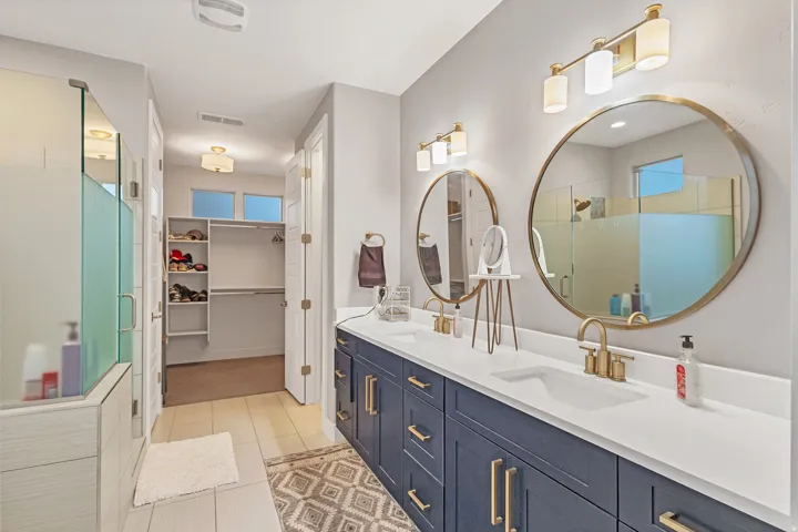 Dual vanity with gold-finish hardware, round framed mirrors, a walk-in closet with built-in shelving, a glass-enclosed shower, and wood-finish flooring