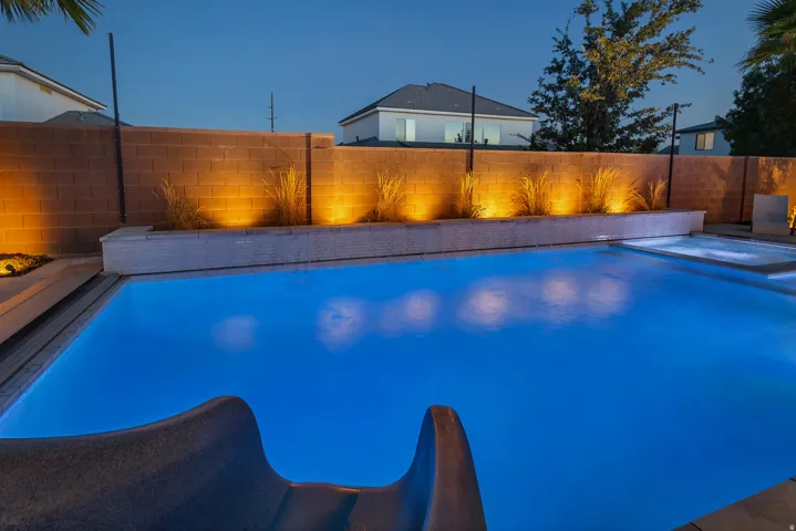 Swimming pool with an integrated spa, a built-in water slide, perimeter accent lighting, and a textured privacy wall