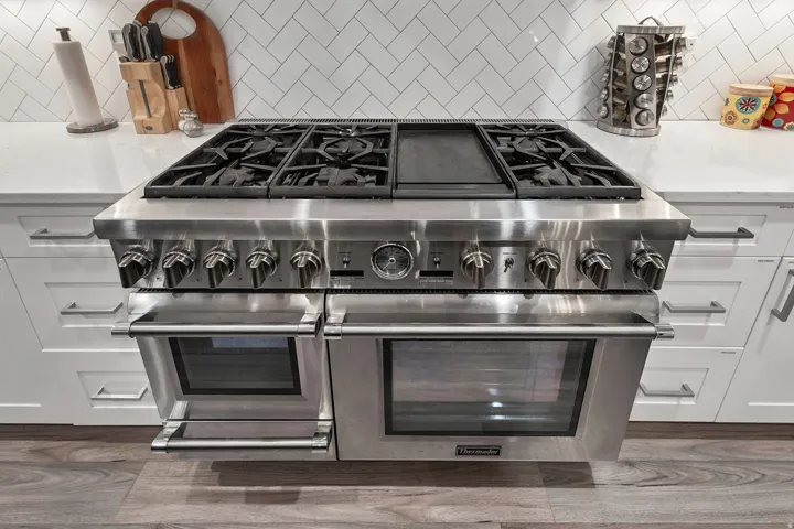 Stainless steel range with multiple burners and integrated griddle, double ovens, white shaker cabinetry, white countertops, and herringbone tile backsplash