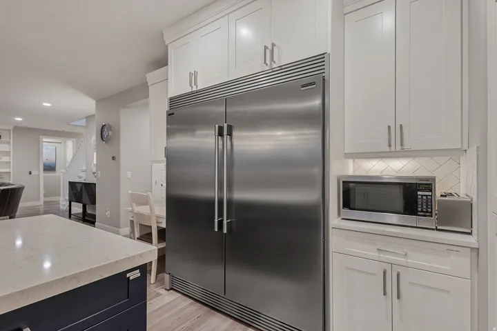 Stainless steel refrigerator, white shaker cabinetry, herringbone tile backsplash, light wood-finish flooring, and a stone countertop