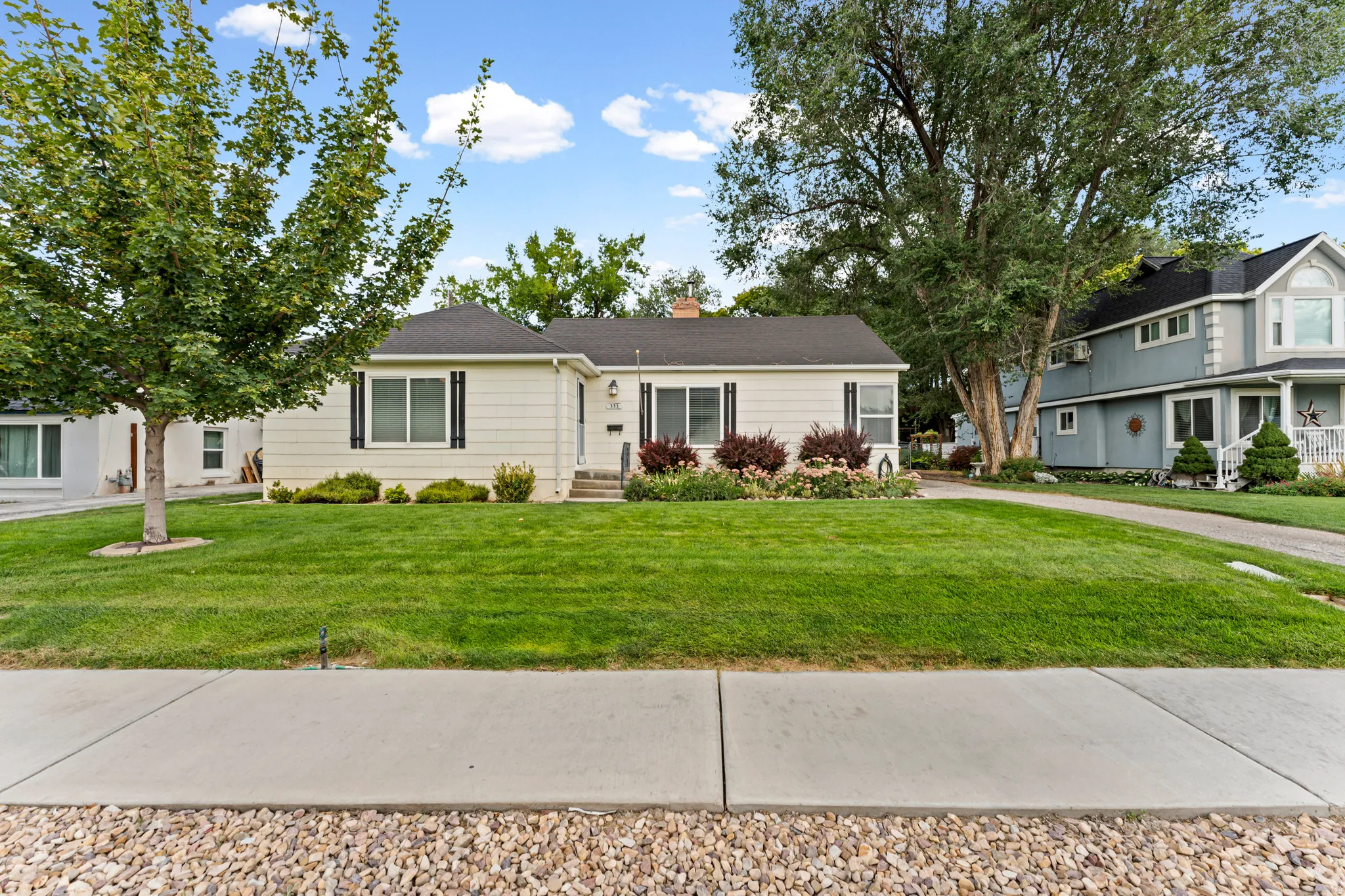 Single-story residence featuring light-colored siding, dark shutters, a front lawn, and mature landscaping