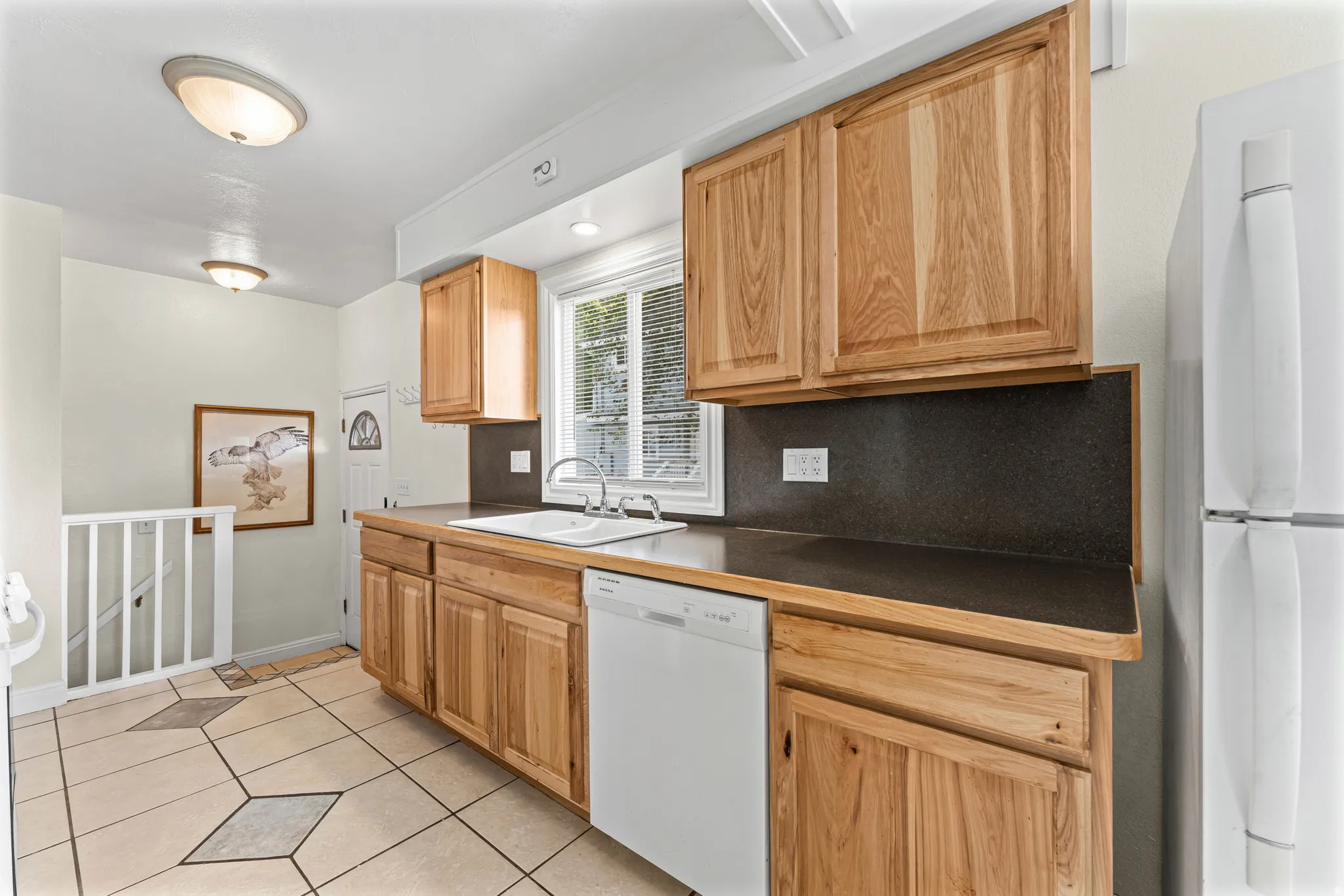 Kitchen with natural wood cabinetry, dark countertops, a white built-in dishwasher, and a double basin sink