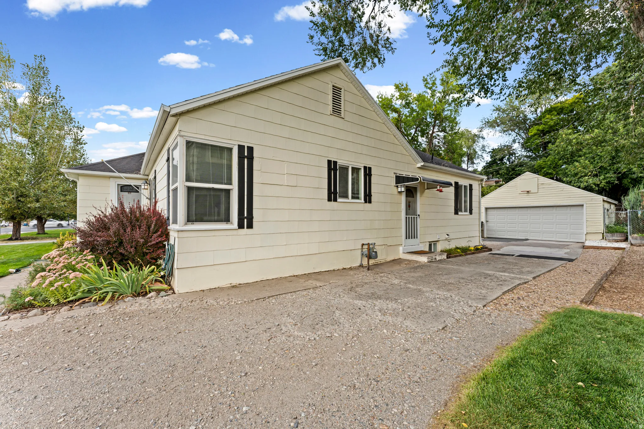 Light-colored siding exterior with black window shutters, a front entry with a small awning, a concrete driveway, a detached garage, and established landscaping