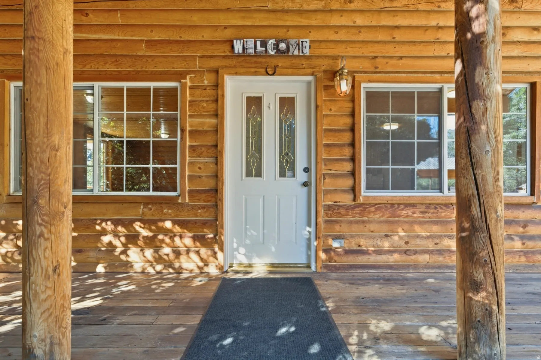 Log cabin exterior featuring a white paneled entry door with decorative glass, two grid-pattern windows, a wall-mounted light fixture, and a wood-plank porch
