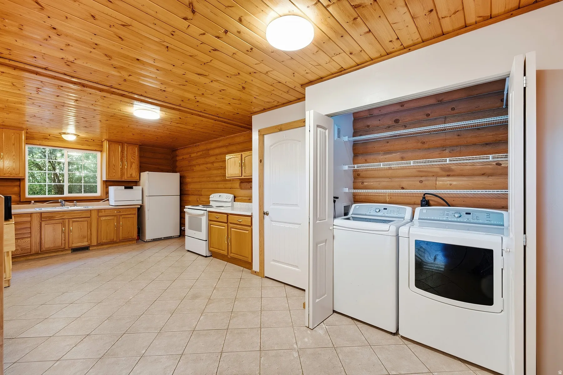 Wood-paneled walls and ceiling, full kitchen with wood cabinetry, tiled flooring, integrated laundry closet with bi-fold doors, and a double-hung window