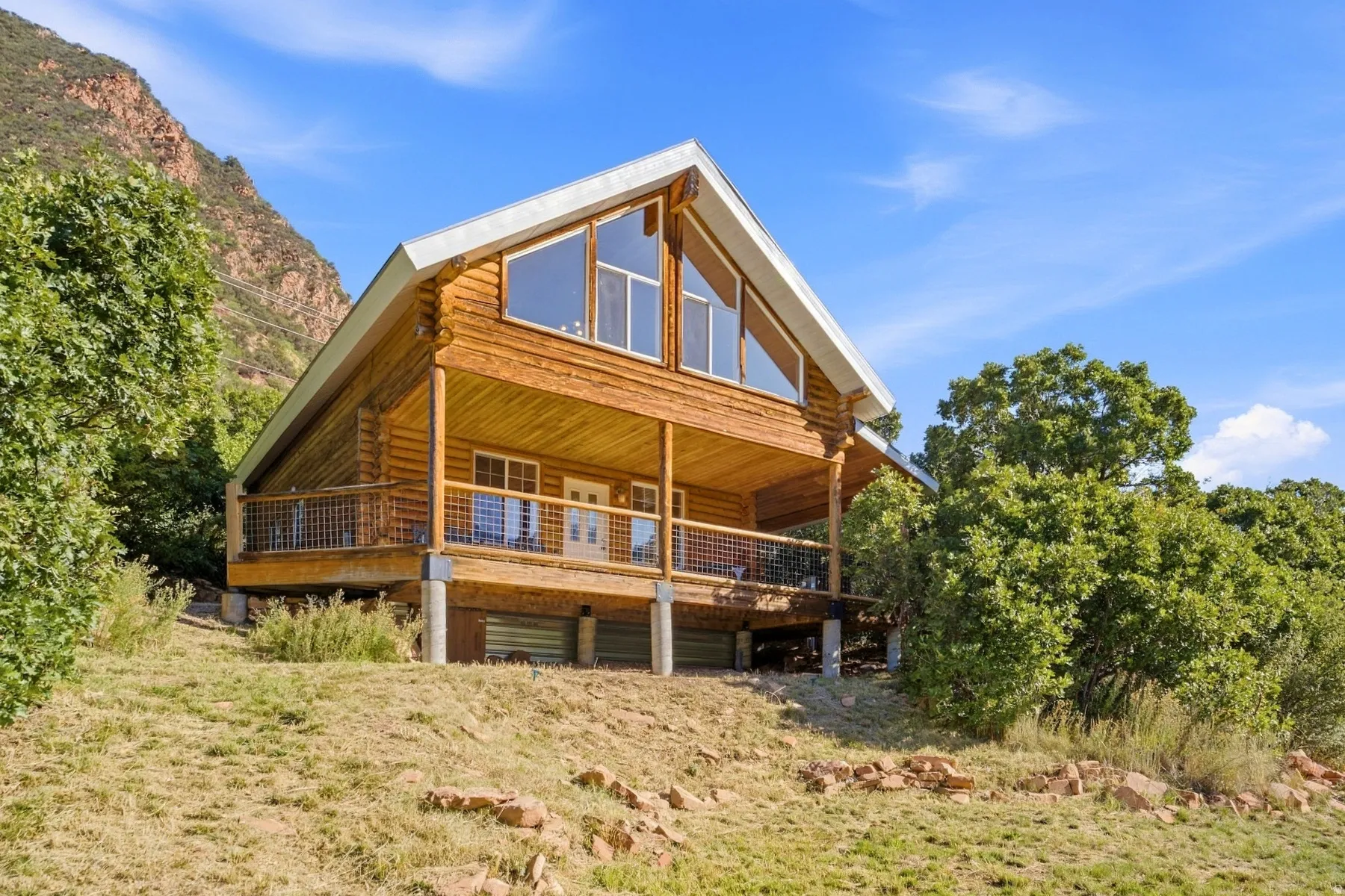 Log cabin exterior with a pitched roof, prominent gable windows, an extended covered deck, and a natural, sloped landscape