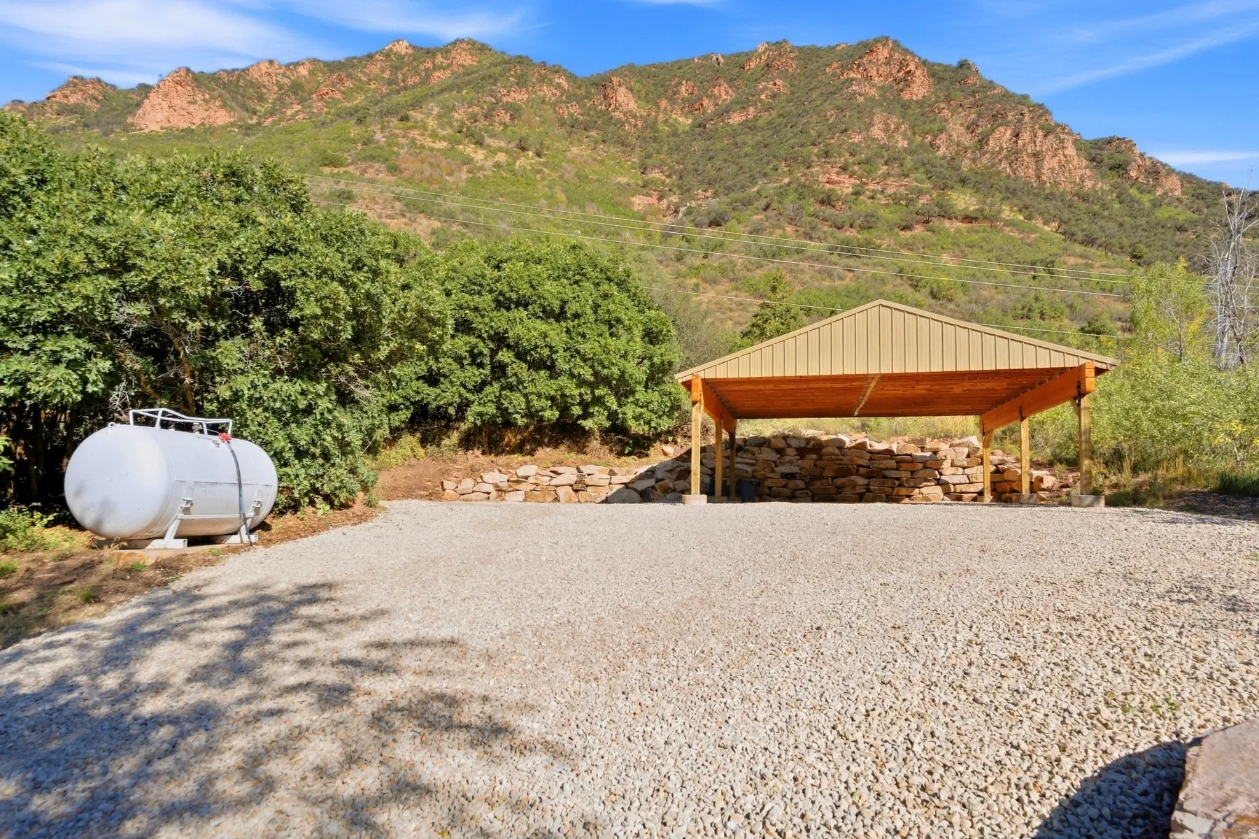 Gravel parking area with a covered carport featuring a metal roof and wood supports, set against a mountainous backdrop