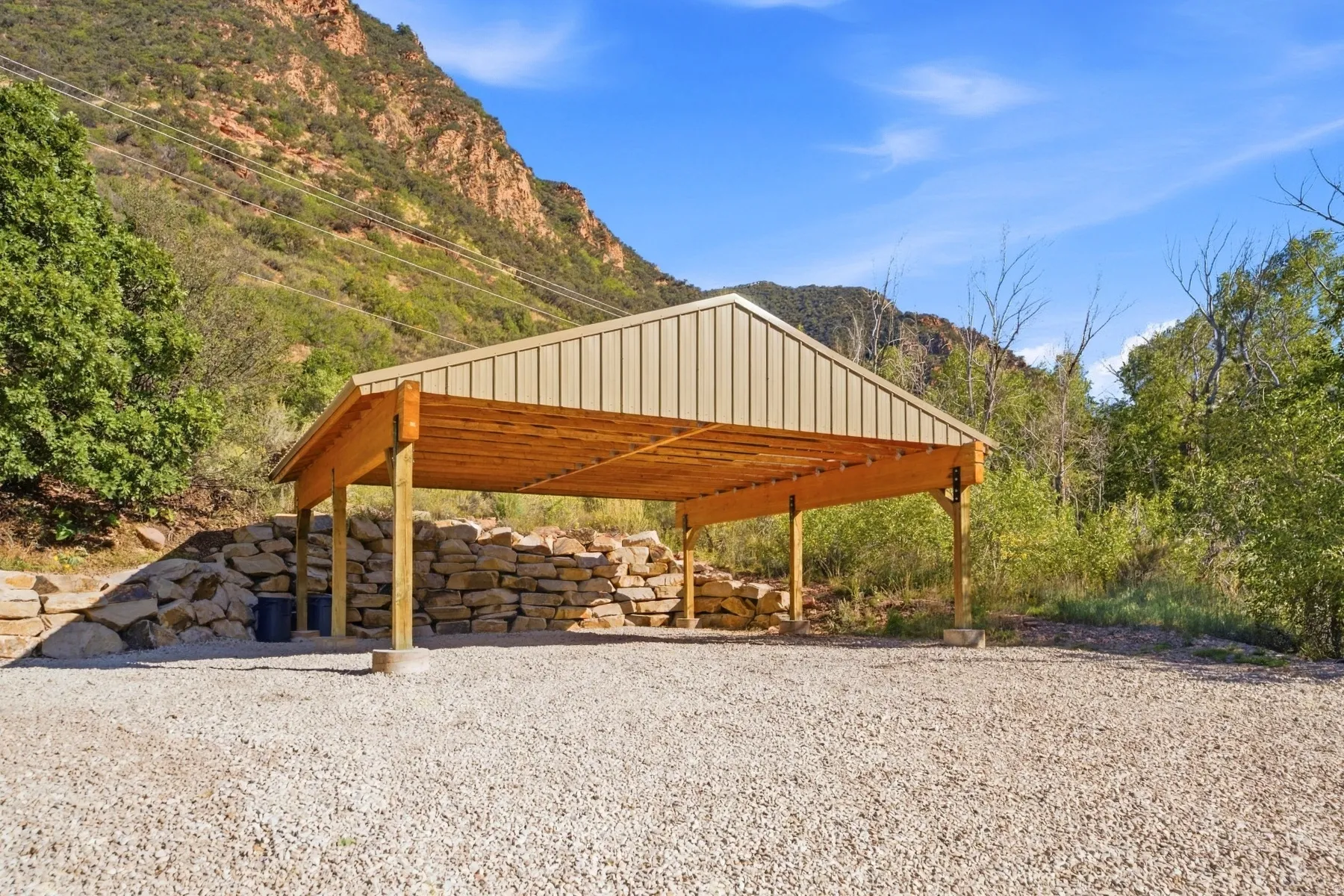 Gravel surface, covered structure, wood beam construction, metal roof, stone retaining wall, mountain backdrop