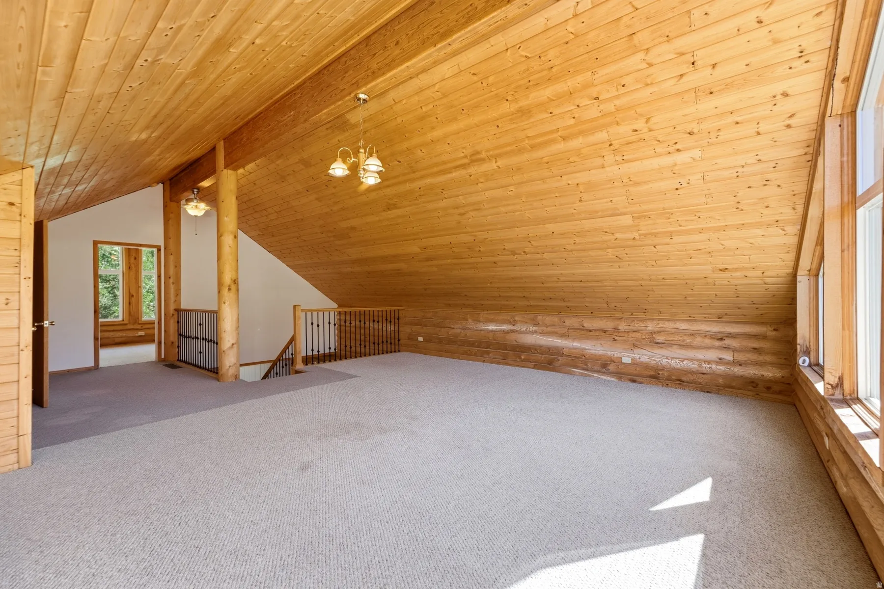 Vaulted wood plank ceiling, exposed structural beam, wood-trimmed windows, light-toned carpeting, and a wooden staircase with iron balusters