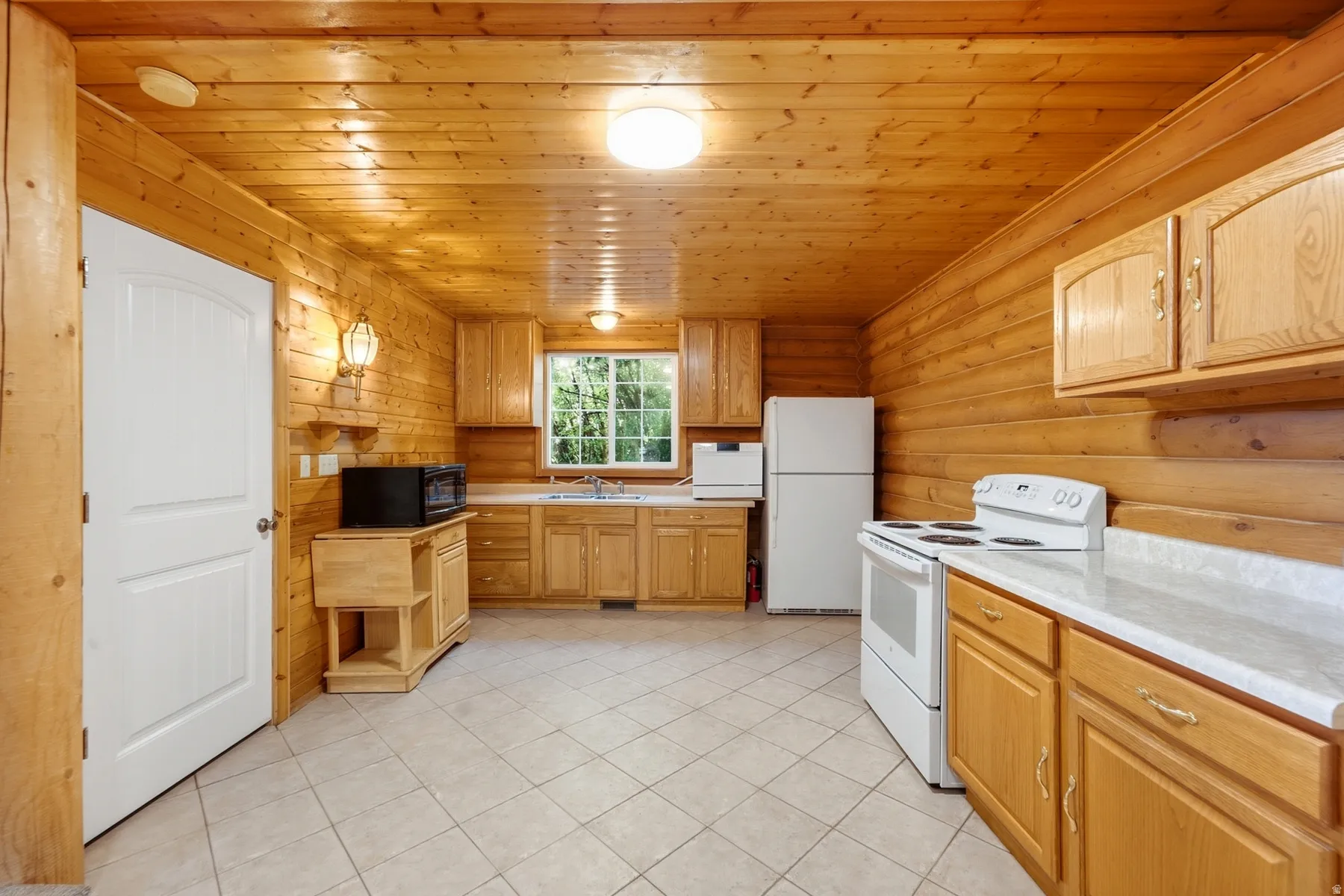 Wood-paneled walls and ceiling, full-height cabinetry, window above the sink, white appliances, and tile flooring