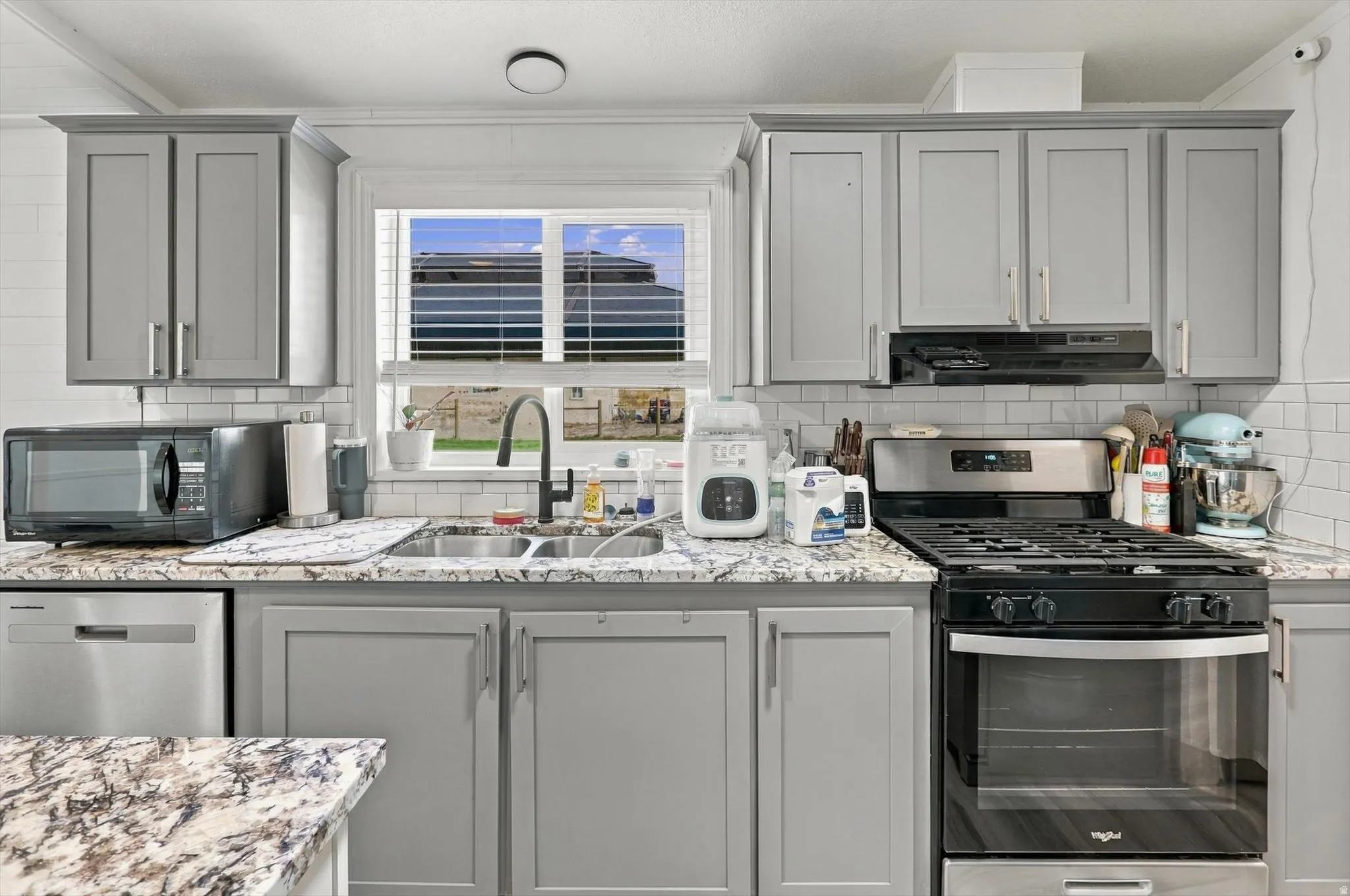 White subway tile backsplash, gray cabinetry, stainless steel dishwasher, dual basin sink with gooseneck faucet, and a stainless steel gas range