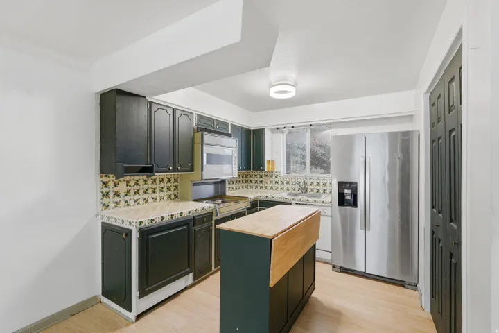 Kitchen featuring dark cabinetry, patterned tile backsplash, light-toned countertops, a stainless steel refrigerator, and wood-finish flooring