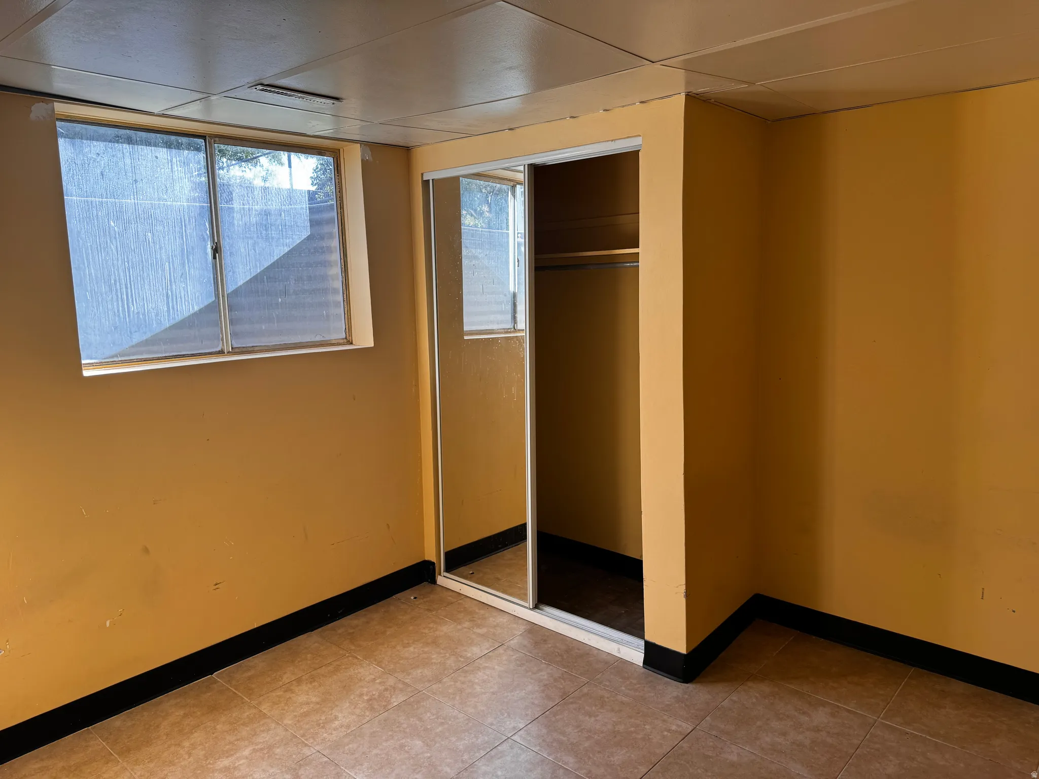 Tile flooring with dark baseboards, complemented by a window with horizontal blinds, and a closet featuring mirrored sliding doors and an interior clothing rod