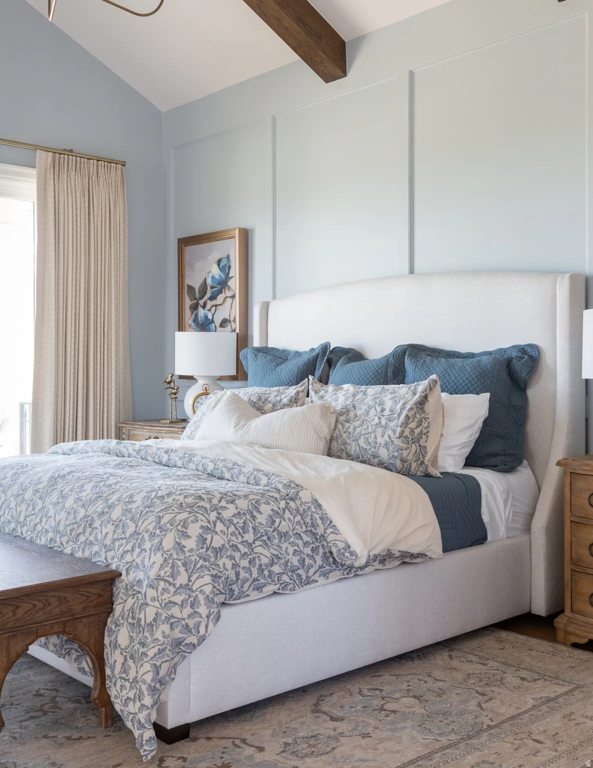 Vaulted ceiling with exposed wood beam, panel molding accent wall, upholstered headboard, wood-finish flooring, and window with drapes