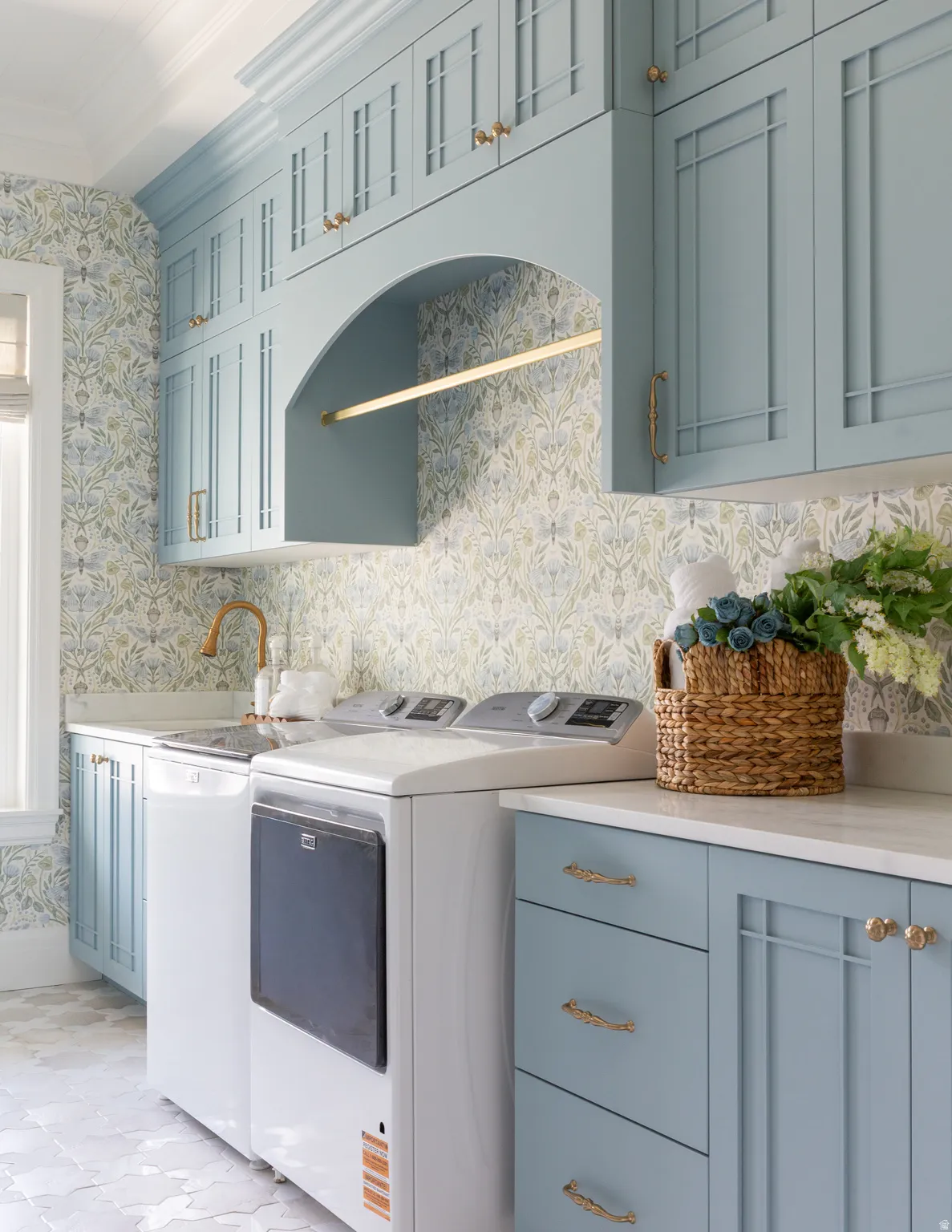 Integrated laundry area featuring light blue cabinetry, built-in shelving, a brass-toned faucet, and patterned wall covering