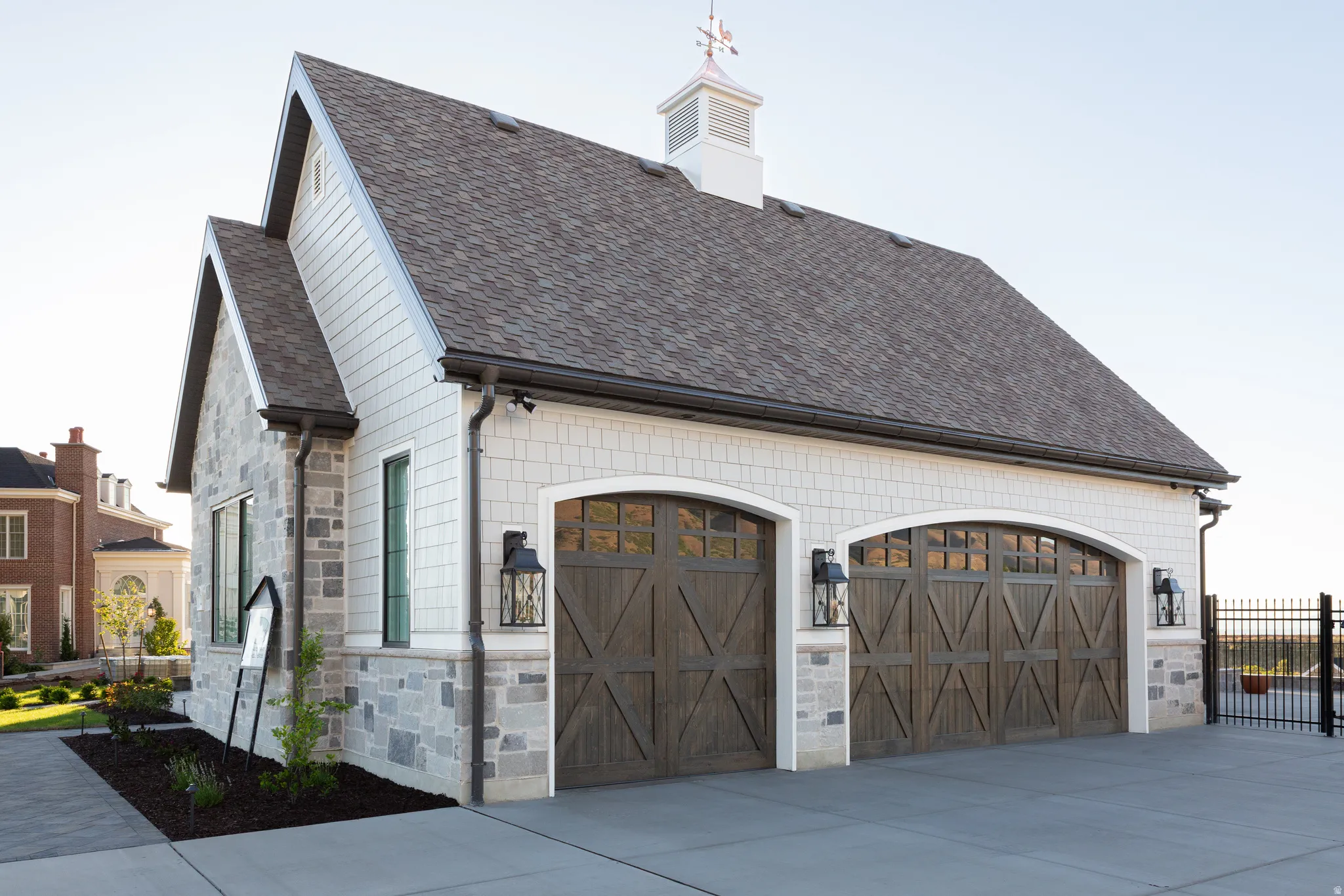 Attached auxiliary building featuring stone and shingle siding, two wood-finish garage doors with arched windows, a cupola with a weathervane, and exterior lanterns