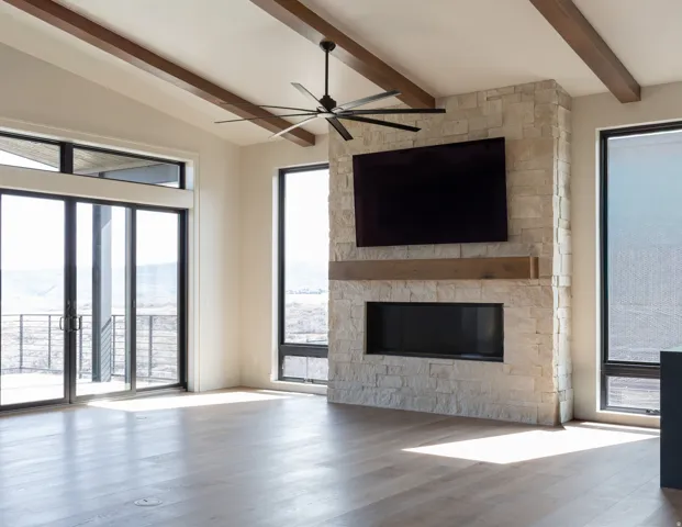 Vaulted ceilings with exposed wood beams, stone-clad fireplace with wood mantle, large format wood-finish flooring, and expansive glass doors