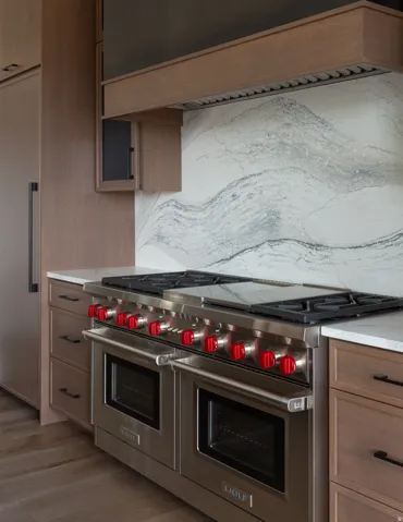 Stainless steel range with double ovens, stone slab backsplash, wood-finish cabinetry, and light-toned countertops