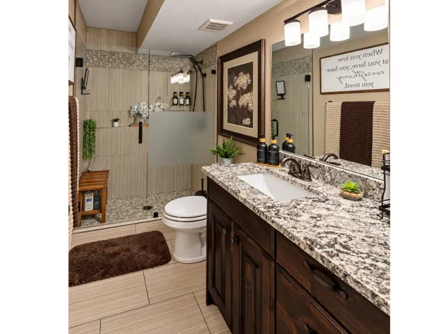 Walk-in shower with a glass enclosure and pebble-tiled floor, vanity with a light-colored countertop, dark wood-finish cabinetry, and a large mirror with integrated lighting