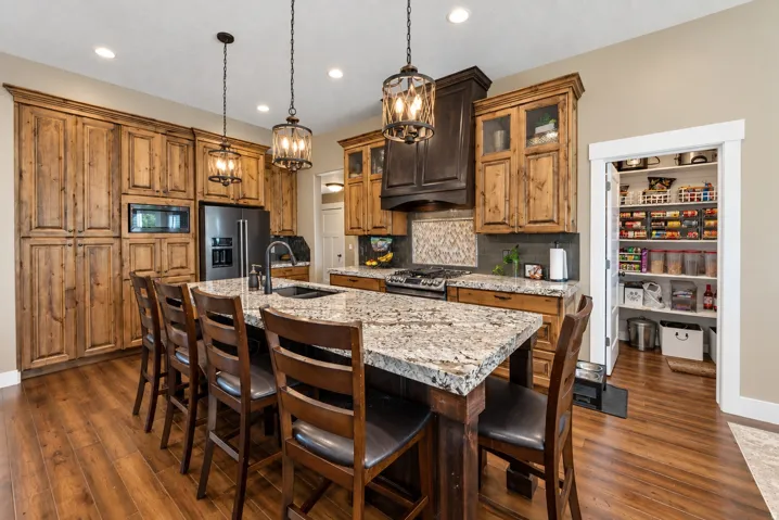 Expansive kitchen island with stone countertop, stainless steel appliances, wood-finish flooring, custom wood cabinetry, and a walk-in pantry