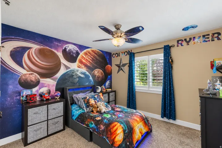 Carpeted room featuring a ceiling fan with integrated lighting, a window with plantation shutters, and a dark-toned built-in storage unit