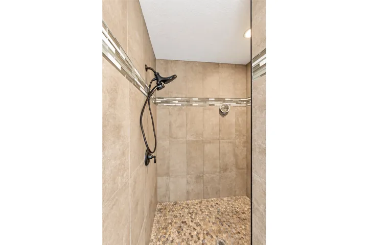 Walk-in shower featuring large format tile walls, a decorative tile accent band, a pebble-finish floor, and a dark bronze shower fixture with a handheld sprayer