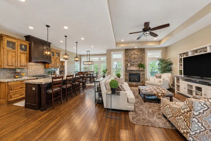 Open-concept living space with wood-finish flooring, a stone fireplace, tray ceiling, kitchen island with seating, and built-in media cabinetry
