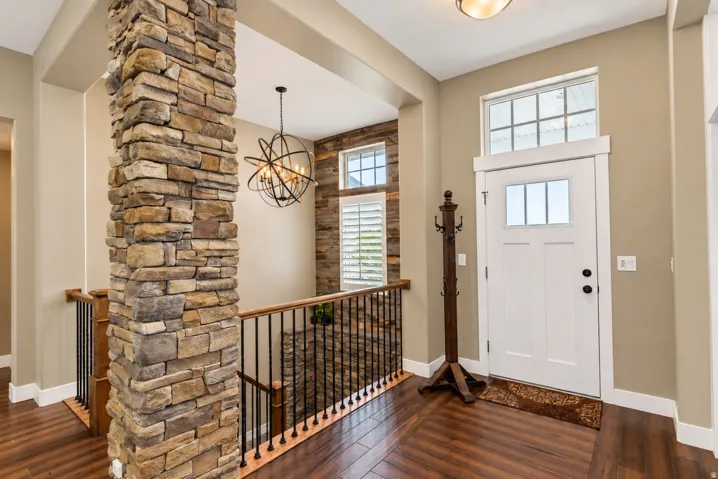 Stone-clad architectural column, wood-finish flooring, white paneled entry door with transom window, wrought iron balusters, and a contemporary spherical chandelier