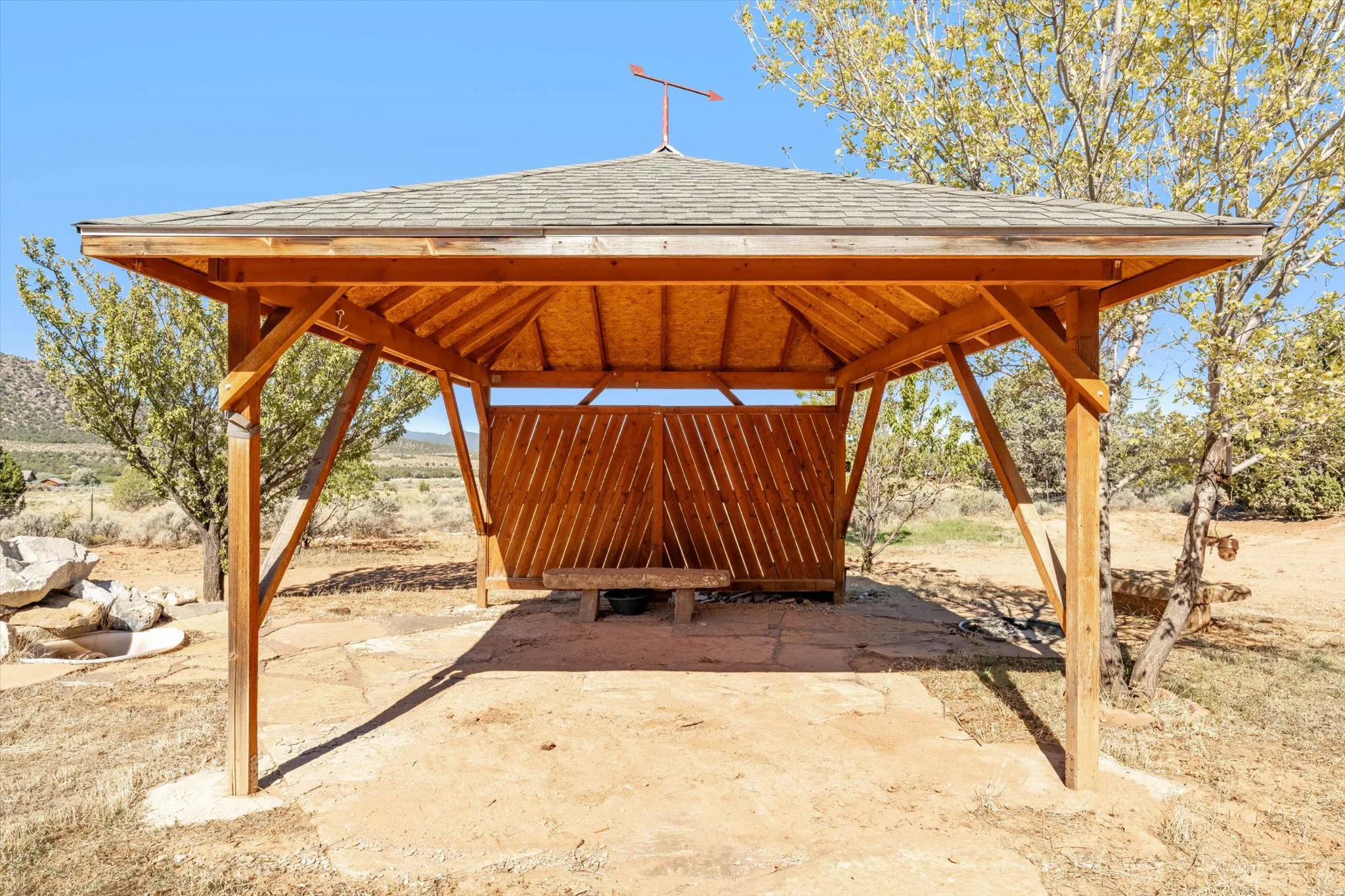 Wood-frame gazebo with a shingled roof, featuring an integrated wooden bench and partial wood-slat walls, set on a flagstone base