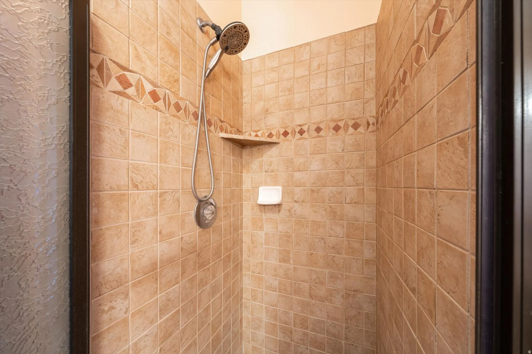 Tiled shower enclosure featuring a decorative accent band, built-in corner shelf, and handheld shower fixture