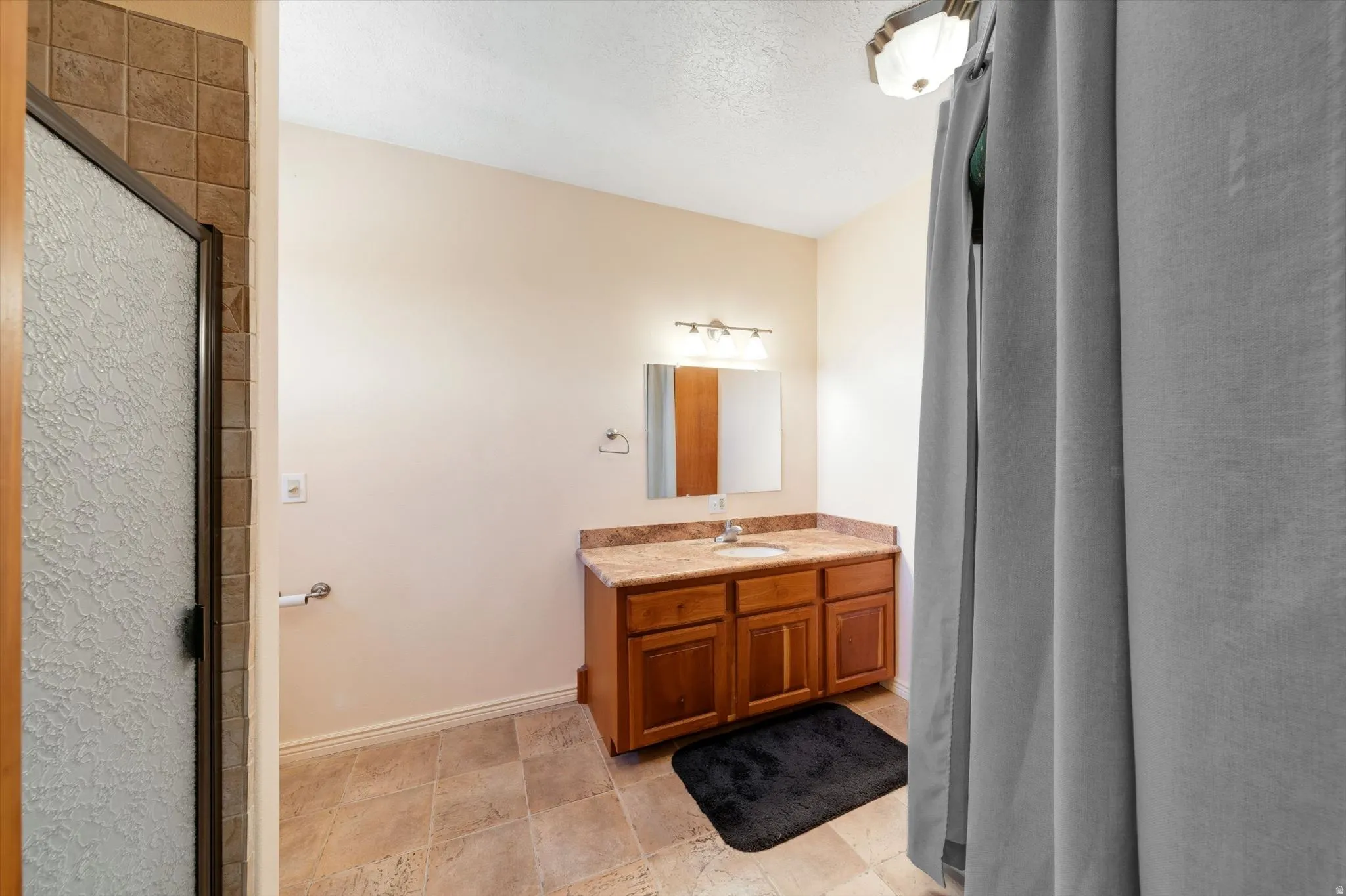 Shower with textured glass door and tile surround, wood-finish vanity with countertop, built-in sink, and wall-mounted mirror, overhead lighting, and tile flooring