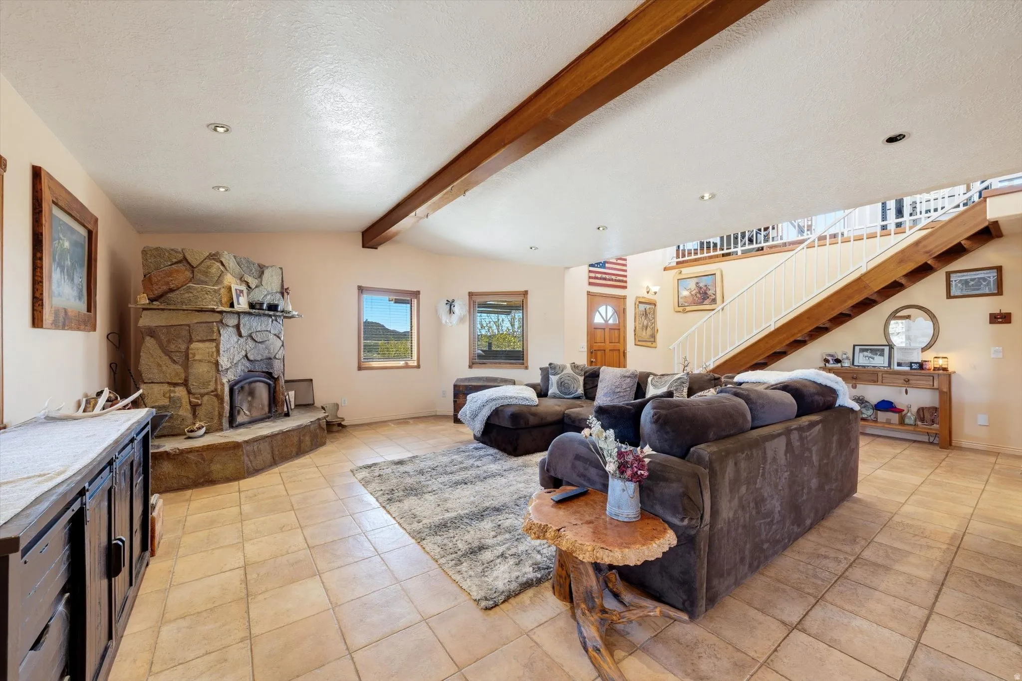 Spacious living area featuring exposed wood beams, a stone fireplace, ceramic tile flooring, recessed lighting, and a prominent wood staircase with white railings