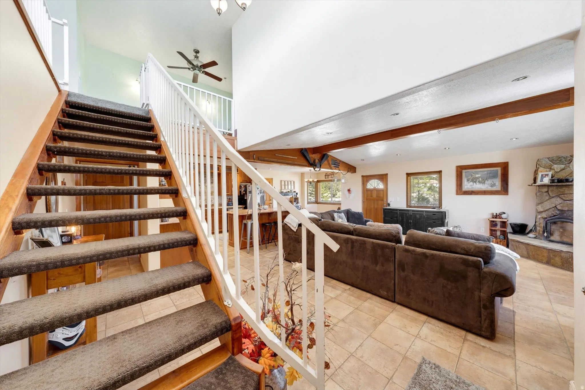 Vaulted ceilings with exposed wood beams, stone-clad fireplace, wood staircase with white railing, ceiling fan, and tile flooring
