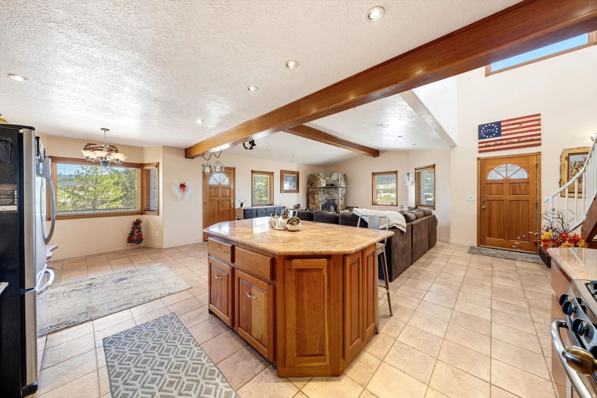 Open-concept living space with exposed wood beams, tile flooring, a central kitchen island with a stone-finish countertop, and a stone fireplace