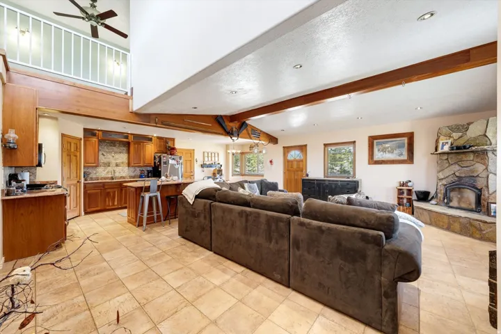 Open-concept living area featuring a stone-faced fireplace, vaulted ceilings with exposed beams, wood-finish cabinetry, and tiled flooring