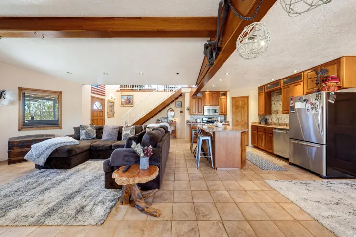 Open-concept living space with tile flooring, exposed wood beams, a kitchen island with seating, stainless steel appliances, and wood cabinetry