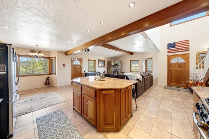 Open-concept living space with exposed wood beams, tile flooring, a central kitchen island with a stone-finish countertop, and a stone fireplace
