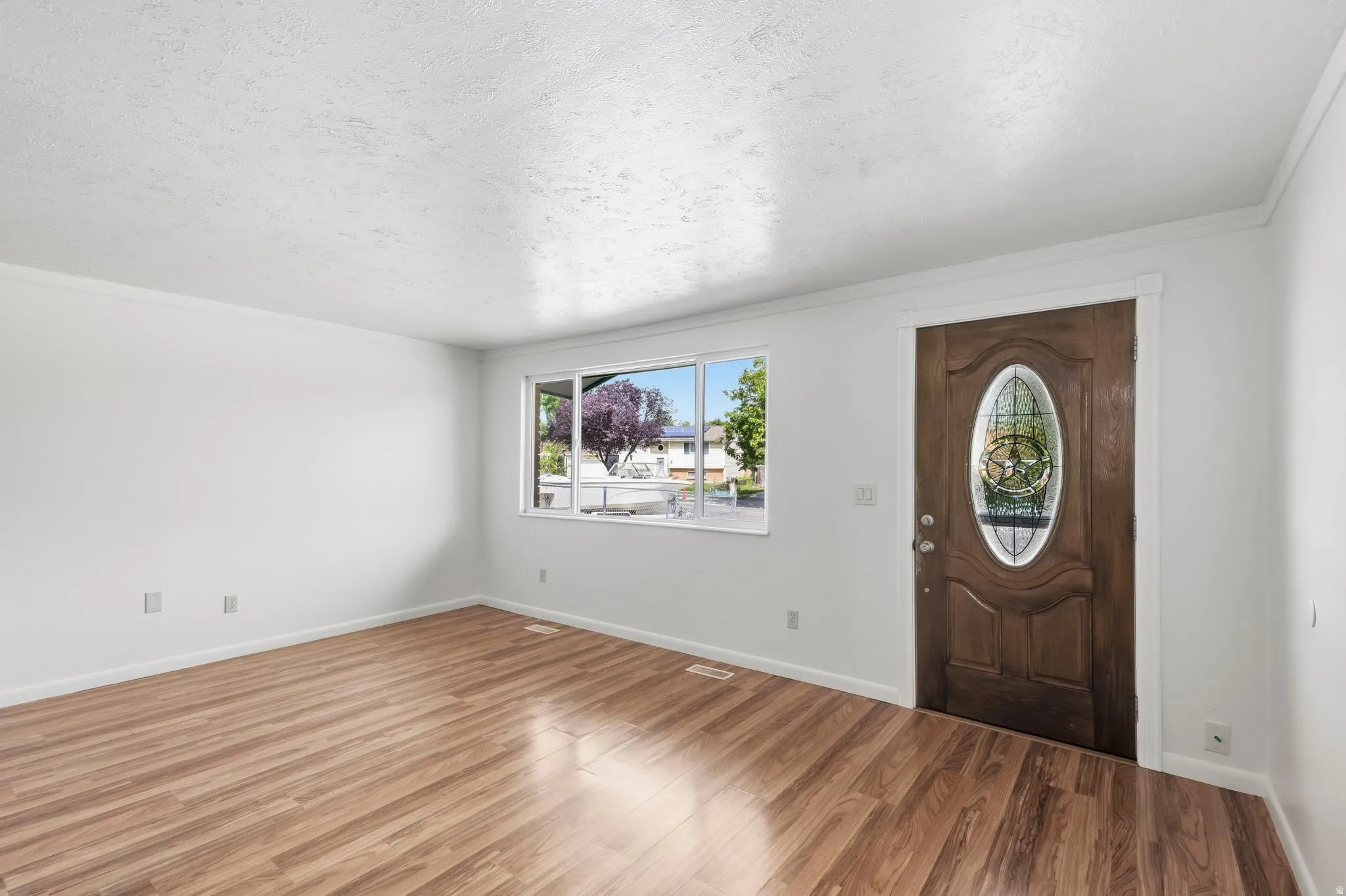 Wood-finish flooring, white painted walls, crown molding, a front entry door with an oval leaded glass insert, and a large window