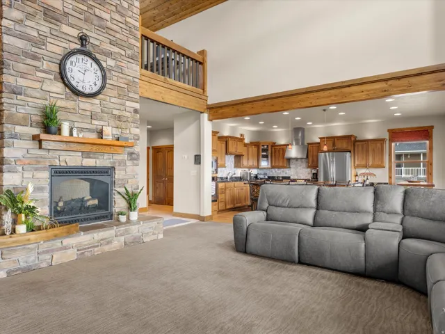 Stone fireplace with a wood mantle, vaulted ceiling with exposed wood beams, open-concept kitchen, wood-finish cabinetry, and stainless steel appliances