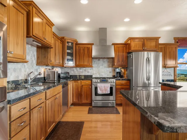 Kitchen featuring wood cabinetry, dark stone countertops, stainless steel appliances, a hexagonal tile backsplash, and wood-finish flooring