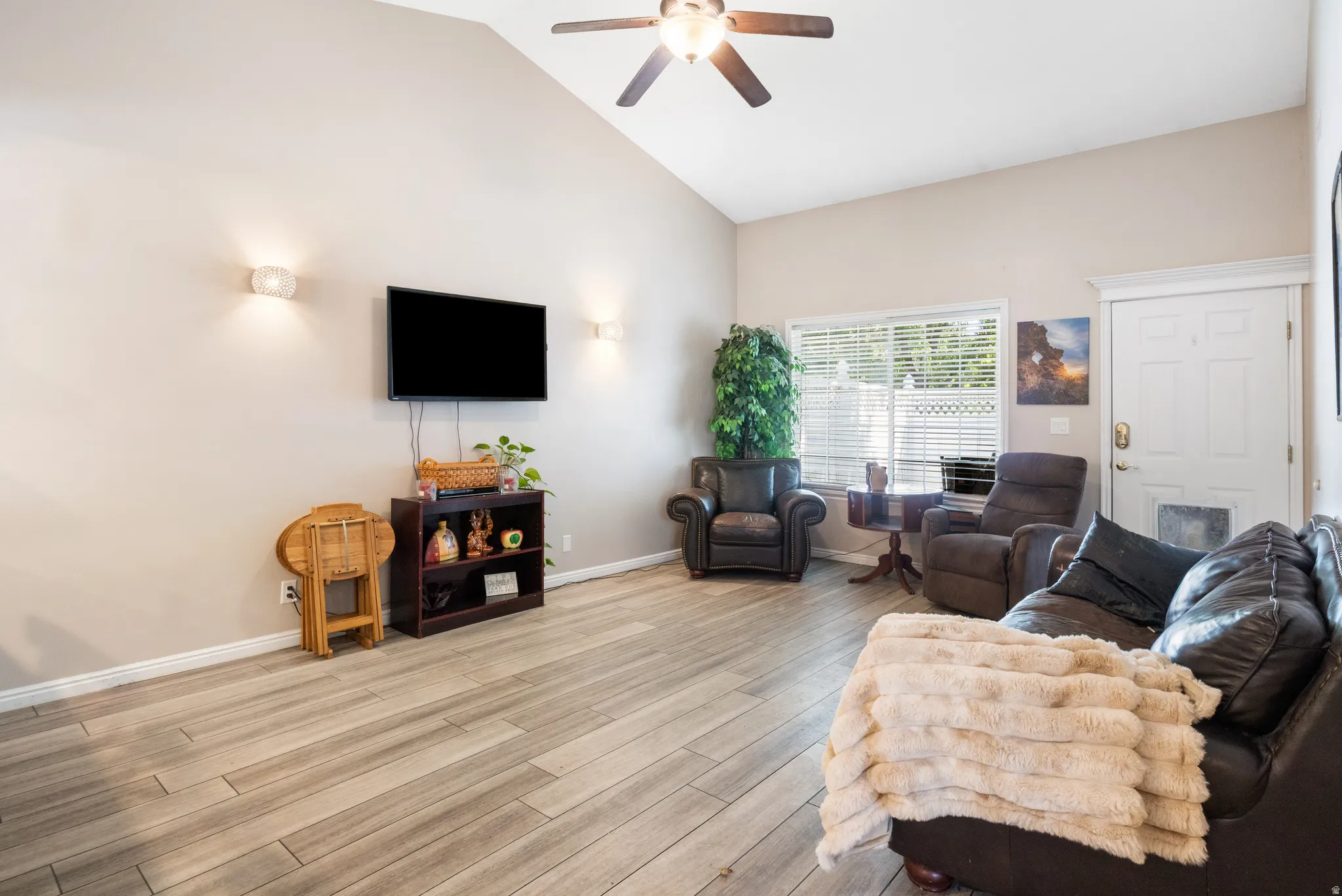 Vaulted ceiling, wood-finish flooring, ceiling fan, wall-mounted sconces, and a bright window with blinds