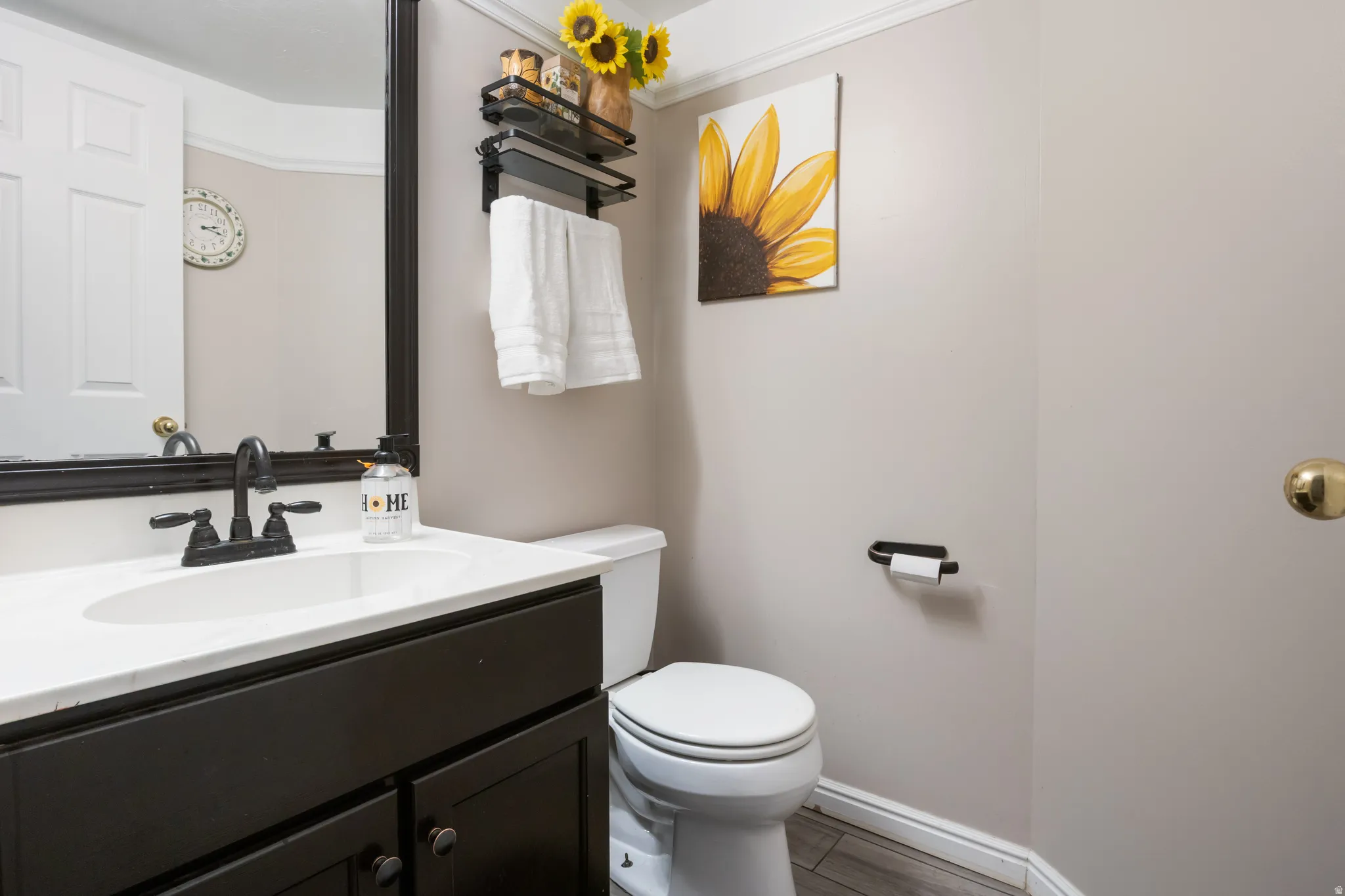 Elegant bathroom featuring a dark vanity with white countertop and integrated sink, matte black faucet and hardware, large framed mirror, toilet with white seat, wood-finish flooring, and soft neutral wall tones accented with sunflower artwork and decorat