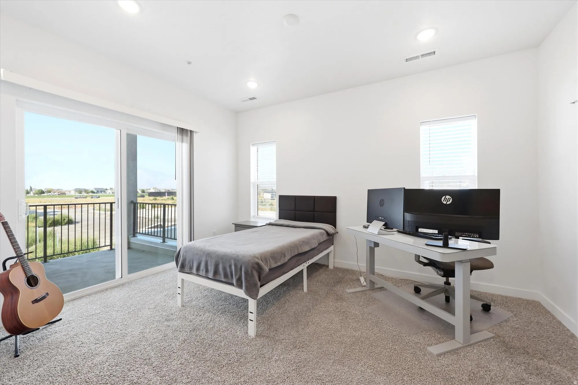 Light-filled room featuring a sliding glass door to a private balcony with black metal railing, recessed ceiling lighting, a window with blinds, and carpeted flooring