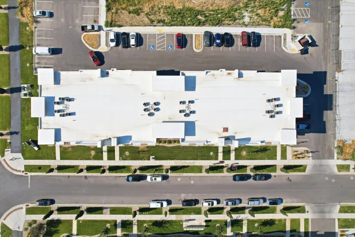 Expansive paved parking areas with designated spaces, concrete sidewalks, green landscaped sections with trees, and a flat roofline